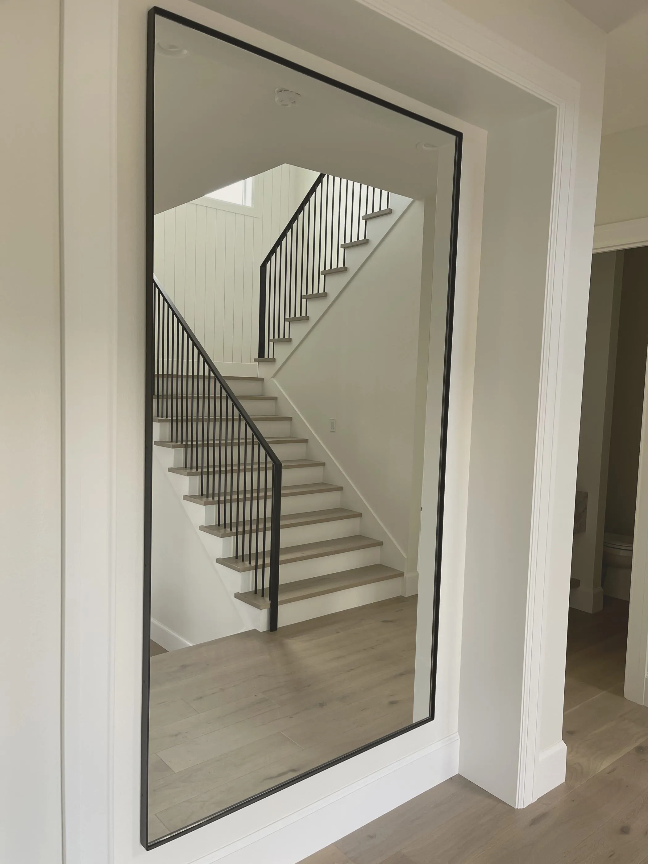 A tall black steel-framed mirror installed in a finished home, reflecting a staircase with wood treads and black balusters.
